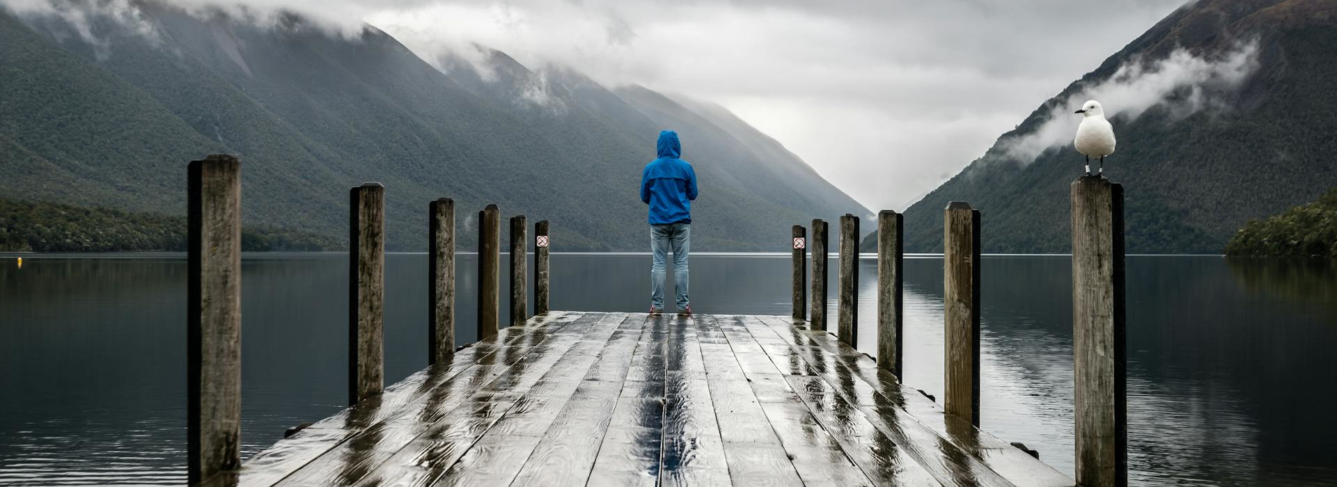 person standing on a lake pier among cloudy mountains