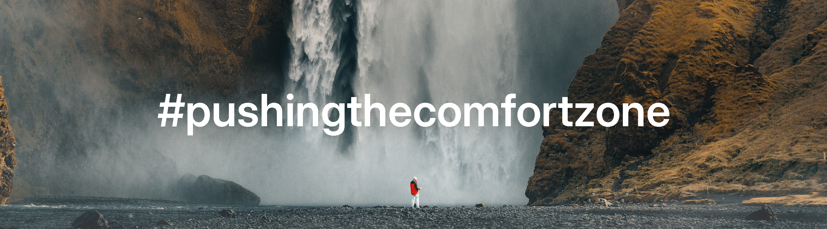 Person standing in front of a massive waterfall in Iceland with '#pushingthecomfortzone' slogan
